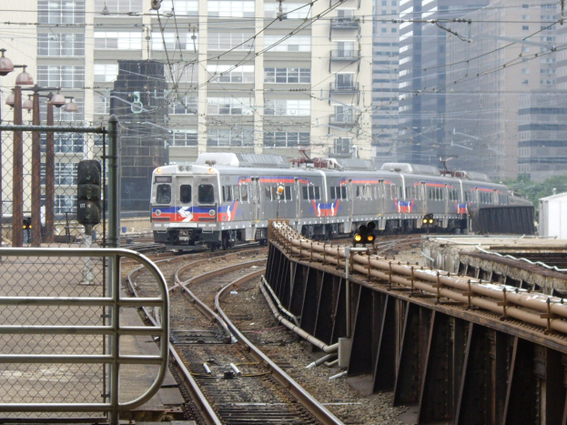 Nahverkehrszug fährt auf Gleisen neben Hochhäusern mit städtischer Infrastruktur und Grünflächen im Hintergrund.