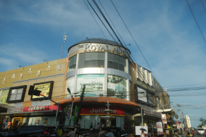 Eine belebte Stadtstraße mit Fahrzeugen, Fußgängern und dem Royal Mall Einkaufszentrum im Blickfeld, mit Bäumen, Pfählen, Drähten und dem Himmel im Hintergrund.