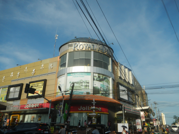 Eine belebte Stadtstraße mit Fahrzeugen, Fußgängern und dem Royal Mall Einkaufszentrum im Blickfeld, mit Bäumen, Pfählen, Drähten und dem Himmel im Hintergrund.