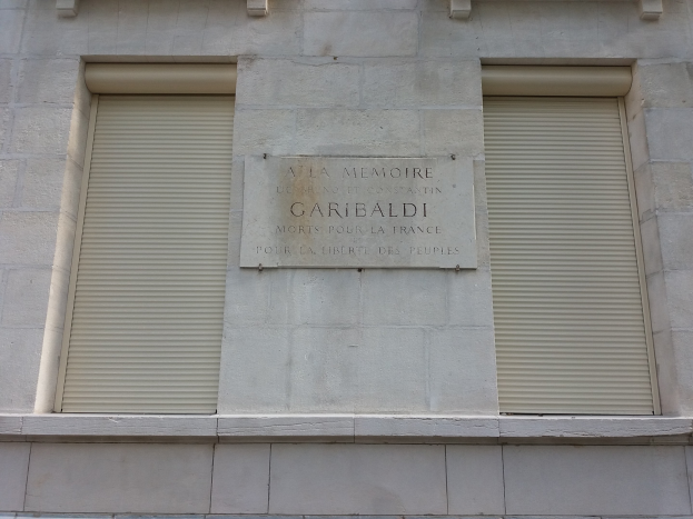 Ein Gebäude mit zwei Fenstern und Läden, das eine Tafel an der Seite trägt, wahrscheinlich ein Denkmal.