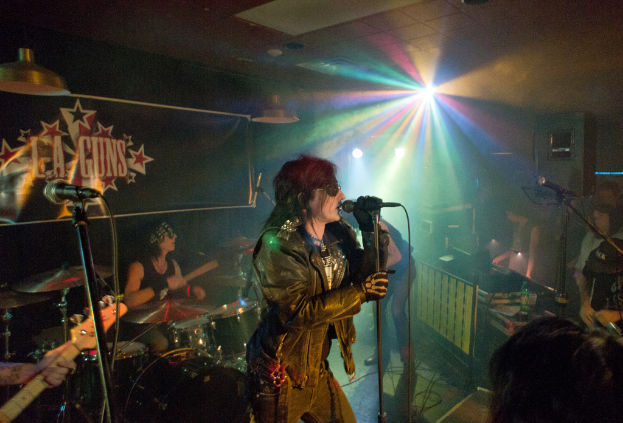 Eine Frau in einer schwarzen Jacke singt in ein Mikrofon auf der Bühne, mit einem Mann in Sonnenbrille und Stirnbinde, der hinter ihr ein Instrument spielt, unter einem Schild, auf dem "LA Guns" steht und bunte Diskolichter leuchten.
