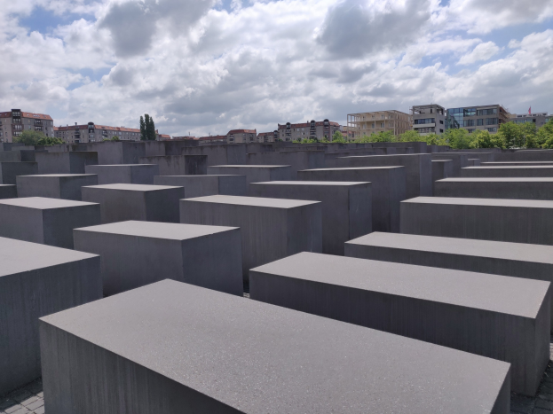 Betonschilder in einem Gittermuster auf dem Holocaust-Mahnmal für die ermordeten Juden Europas in Berlin, mit Bäumen und Gebäuden im Hintergrund unter einem bewölkten Himmel.