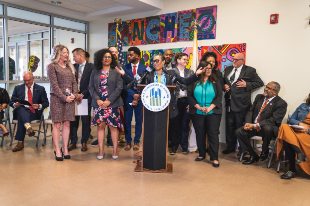 Gruppe von Menschen vor einem Podium mit einem Mikrofon und einer Tafel mit Text, einige sitzen auf Stühlen und andere stehen, mit Flaggen, einer Tafel, einem Feuerlöscher und Deckenleuchten an der Rückwand.