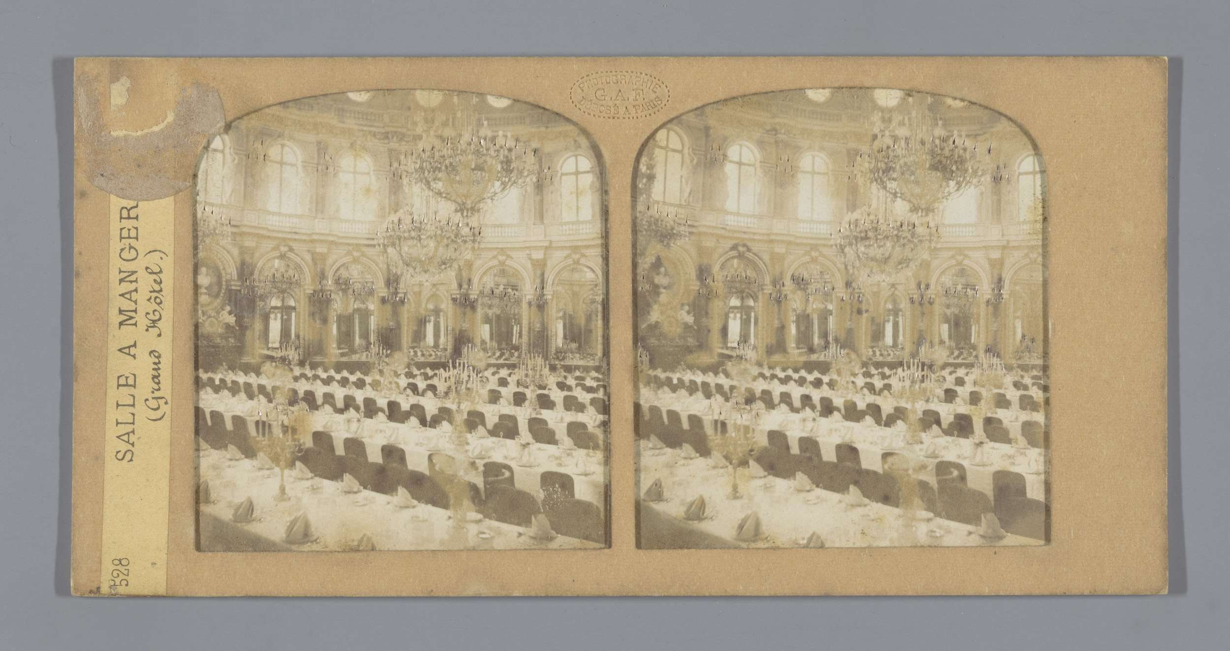 Schwarz-weiß-Foto eines prächtigen Hotel-Speisesaals mit Tischen, Stühlen, Kronleuchtern und Fenstern, mit einer Tafel links mit der Aufschrift "Sale a Manger, Grand Hotel".