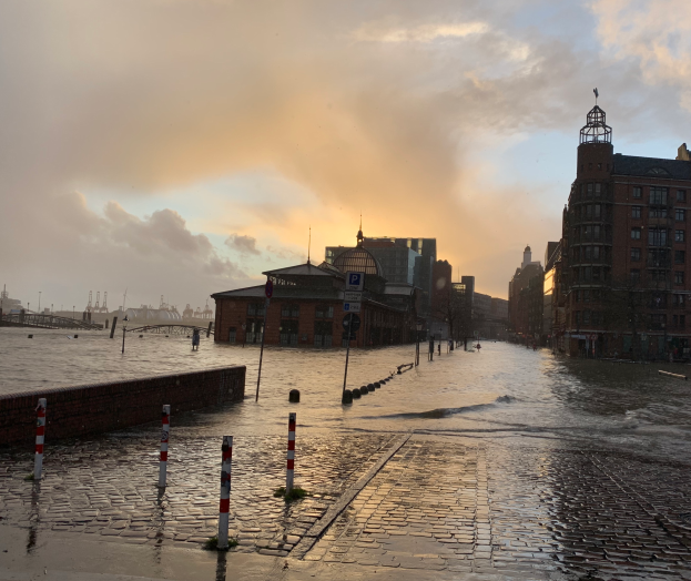 Eine überflutete Straße in Hamburg, Deutschland, mit Wasser bedeckt, Pfählen, Schildern, Gebäuden mit Fenstern, einer Brücke und einem bewölkten Himmel im Hintergrund.