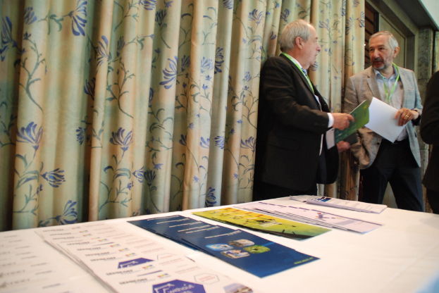 Männer mit Mänteln und Ausweisen halten Bücher, mit einem Vorhang im Hintergrund und Papieren auf einem Tisch im Vordergrund.