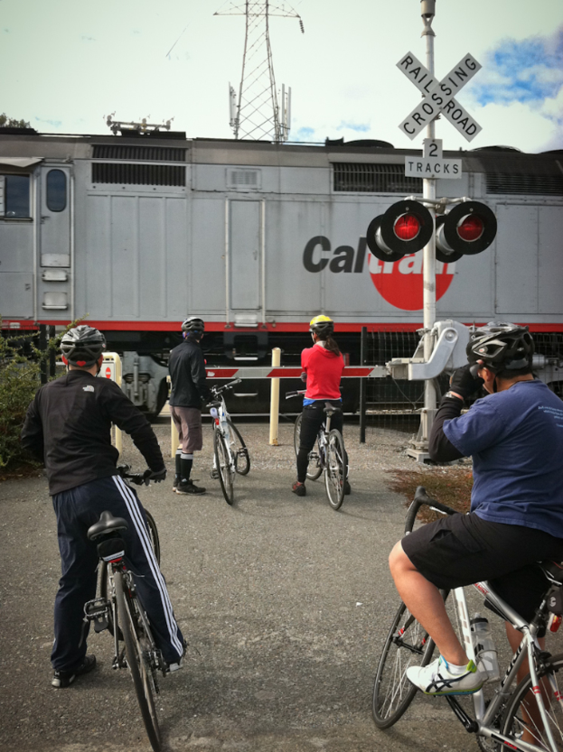 Vier Menschen mit Fahrrädern stehen auf einer Straße in der Nähe eines Bahnsignals und eines Tors.