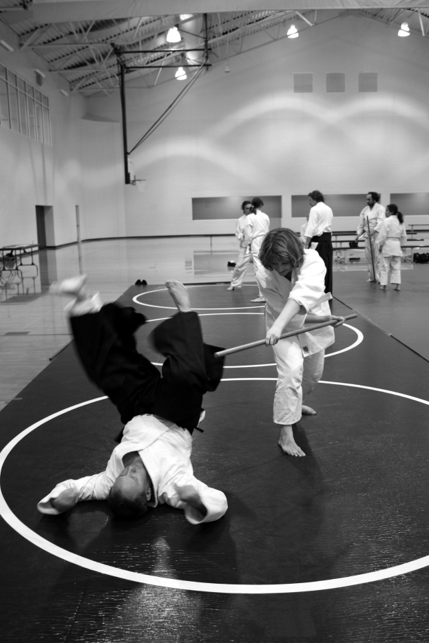 Gruppe von Menschen, die Karate in einem Fitnessstudio ausüben, zwei Personen kämpfen mit Stöcken in der Mitte, andere stehen drumherum, Wand mit Lampen im Hintergrund.