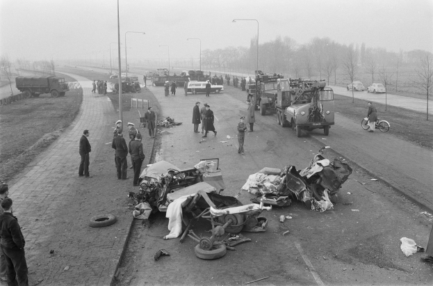 Schwarze und Weiße Szenerie eines Autounfalls am Straßenrand mit mehreren Fahrzeugen, darunter ein Geländefahrzeug, und einer Gruppe von Menschen, die darum herumstehen, mit Laternenpfählen, Bäumen und Himmel im Hintergrund.