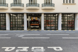 Eine Feuerwache mit einem Feuerwehrauto davor, mit Fenstern, Geländern, Säulen und Glastüren, mit einer Straße unten.