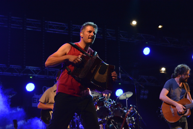 Drei Menschen auf einer B├╝hne, die musikalische Instrumente spielen, mit einer Person, die eine Brille tr├Ągt, und Scheinwerfern und St├Ąben im Hintergrund.