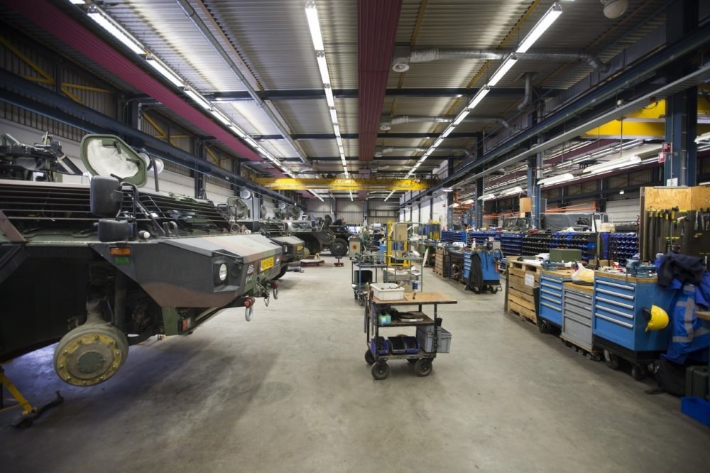 Großes Lager mit Maschinen, Fahrzeugen links, Wagen mit Gegenständen, Holzboxen und Deckenbeleuchtung.