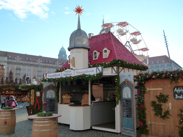 Ein kleines Gebäude mit einem roten Dach und einem Riesenrad davor, umgeben von Menschen und festlichen Dekorationen bei einem Outdoor-Event.