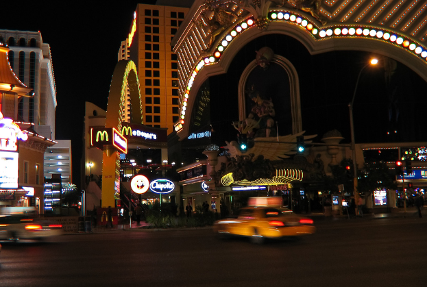 Eine nächtliche Stadtansicht mit Gebäuden, Ampeln, einer Straßenlaterne, einer Statue, Bannern und Lichtern vor einem dunklen Himmel.