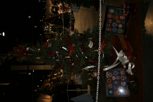 Ein Weihnachtsbaum mit Geschenken, Rahmen und Spielzeug auf einer Plattform, mit Menschen, zusätzlichen Weihnachtsbäumen, Ketten, Pfählen und Lichtern im Hintergrund.