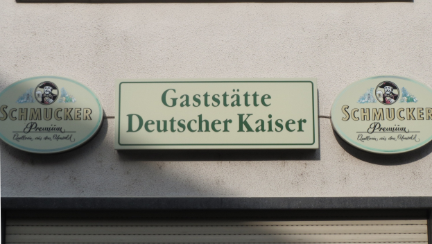 Gebäude mit drei wandmontierten Tafeln, auf denen der Text "Gaststätte Deutscher Kaiser Schmucker" steht, und einem Rollladen unten.