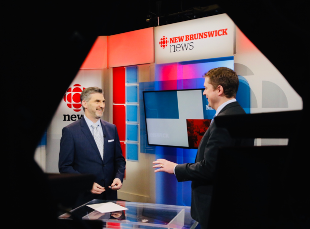 Zwei Männer in Blazern und Krawatten unterhalten sich vor einem Glastisch mit Papieren, im Hintergrund ein Fernseher mit der Aufschrift "New Brunswick News".