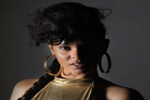 Eine Frau in einem goldenen Kleid und großen Ohrringen steht selbstbewusst vor einer Wand, ihr glattes Bob-Haar und natürliches Make-up unterstreichen ihren entschlossenen Ausdruck, während sie direkt in die Kamera schaut.