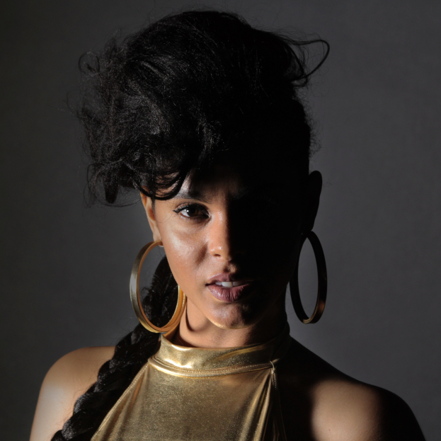 Eine Frau in einem goldenen Kleid und großen Ohrringen steht selbstbewusst vor einer Wand, ihr glattes Bob-Haar und natürliches Make-up unterstreichen ihren entschlossenen Ausdruck, während sie direkt in die Kamera schaut.