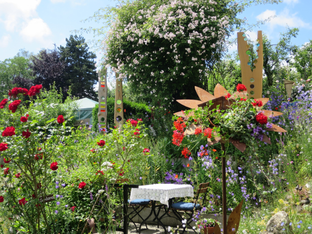 Ein Garten mit bunten Blumen und Pflanzen, der einen zentralen Tisch mit einer Decke und Stühlen zeigt, umgeben von Bäumen unter einem bewölkten Himmel auf dem Chelsea Flower Show.