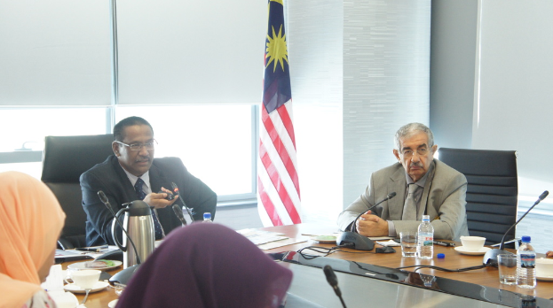 Eine Gruppe von Menschen, darunter Männer in Anzügen und zwei Frauen in Burkas, sitzt um einen Tisch mit Wasserflaschen, Mikrofon, Tassen und einem Teekessel, mit einem Fenster und einer Flagge im Hintergrund.