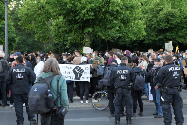 Eine große Gruppe von Menschen steht auf der Straße, einige halten Schilder, mit einem Fahrrad im Vordergrund und Bäumen und einem Mast im Hintergrund, bei einer Black-Lives-Matter-Demonstration in Berlin.