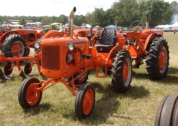 Mehrere orangen Traktoren sind in einem Garten mit Gras am Boden, Zelten in Grün, Blau und Weiß, Bäumen, Gebäuden und Fahrzeugen im Hintergrund und zwei Männern auf der rechten Seite unter einem klaren Himmel geparkt.