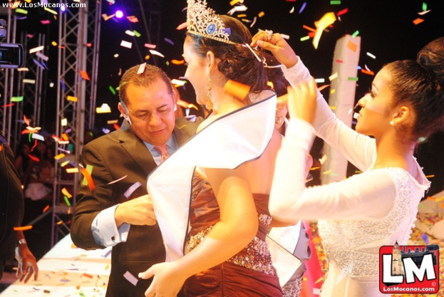 Ein Mädchen auf der Bühne bei einer Miss World Show mit einer Krone, dahinter ein Mädchen, das die Krone anpasst, und ein Mann in einem schwarzen Anzug, der vorne die Hand schüttelt, vor einem dunklen Hintergrund mit Metallrahmen.