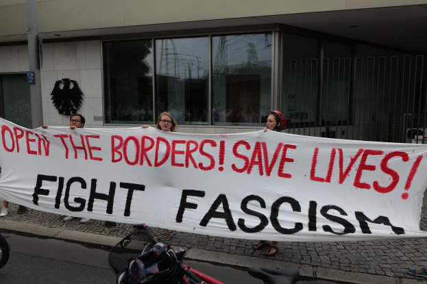 Eine Gruppe von Menschen mit einem Transparent mit der Aufschrift "Öffnet die Grenzen, rettet Leben, bekämpft Faschismus" vor einem Gebäude mit Glasfenstern und einem Pfahl, mit einem Fahrrad und einer Tasche auf der Straße.