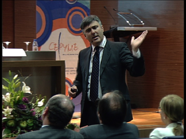 Ein Mann in einem schwarzen Blazer steht an einem Podium mit Mikrofonen, spricht in ein Handmikrofon, mit einer Blumenanordnung und einem Tisch mit einem Buch und einer Wasserflasche in der Nähe, während drei Personen auf Stühlen vor ihm sitzen, gegen den Hintergrund einer braunen Wand und einer bunten Tafel mit Text.