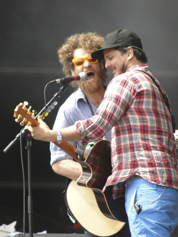Zwei Männer auf der Bühne, einer in rot-blau kariertem Hemd und blauen Hosen, der andere mit Brille und schwarzer Mütze, beide singen in Mikrofone und spielen Gitarren.