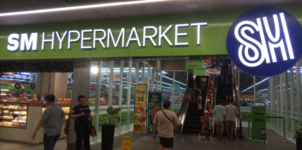 A group of people walking in front of a brightly lit 'SM Hypermarket' store with ceiling lights, text boards, and houseplants visible inside.