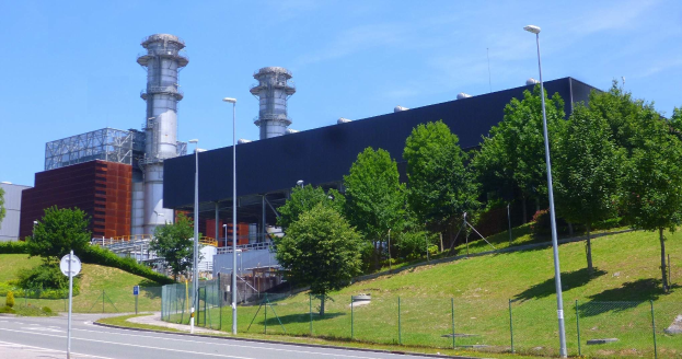 Großes Industriegebäude mit vielen Fenstern, als Kraftwerk identifiziert, umgeben von Straßeninfrastruktur, Fahrzeugen, Grünflächen und einem bewölkten Himmel.
