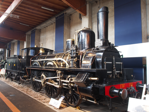 Eine große, alte Dampflokomotive in einem Museums-Setting umgeben von Eisenbahnschienen, Informations-Tafeln und anderen Ausstellungsstücken, mit einer beleuchteten Decke und Wand im Hintergrund.