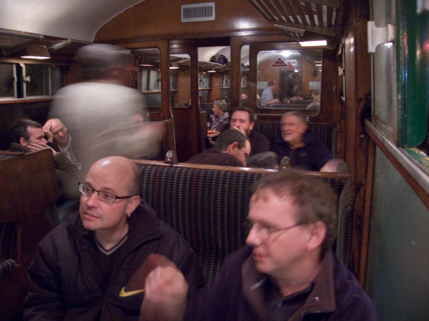 Zwei Männer sitzen auf einem Sitz in einem Zug, mit einer Gruppe von Menschen, die dahinter sitzen, einem Eingang zwischen Abteilen oben und einem Fenster rechts.