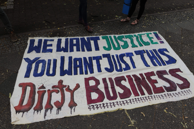 Ein Banner auf der Straße mit der Aufschrift "Wir wollen Gerechtigkeit, ihr wollt nur diesen schmutzigen Deal" und zwei Personen oben drauf, eine hält ein blaues Objekt.
