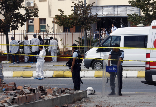 Eine Gruppe von Polizeibeamten steht vor einem Gebäude mit Fenstern, umgeben von Fahrzeugen, einem Zaun, Bäumen und Pflanzen, mit Ziegelsteinen und einem Helm auf dem Boden, wahrscheinlich nach einer Bombenexplosion in Mexiko-Stadt.