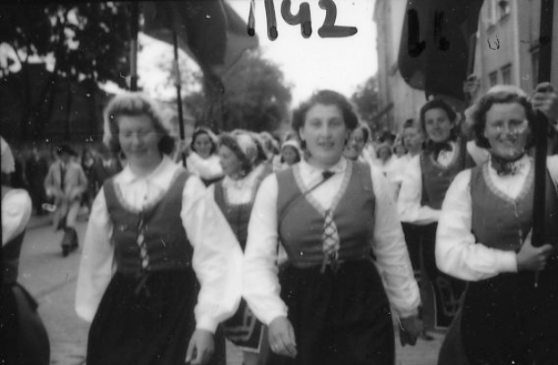 Eine Gruppe von Frauen in traditioneller bayrischer Tracht spaziert strahlend durch eine Straße, umgeben von Bäumen und Gebäuden, dargestellt in Schwarz-Weiß.