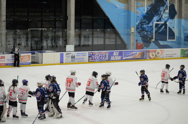 Gruppe junger Menschen, die Eis hockey auf einer Indoor-Eisfläche spielen, mit Helmen und Schlägern ausgestattet und mit einer Person auf der linken Seite und Bannern, Säulen und Glaswänden im Hintergrund.