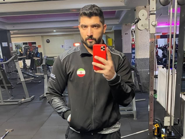 Ein Mann macht ein Selfie in einem Fitnessstudio, umgeben von Geräten und unter Deckenbeleuchtung, mit einem Handy in der Hand.