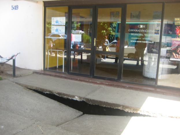 Eine gebrochene Fläche im Vordergrund mit einer Glastür, Wand und Stange im Hintergrund, die sich bewegende Fahrzeuge und einige stehende Personen spiegeln.