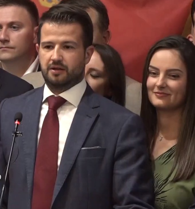 Ein Mann im Anzug spricht in ein Mikrofon an einem Rednerpult, mit einem Banner hinter ihm, zu einer Menge.