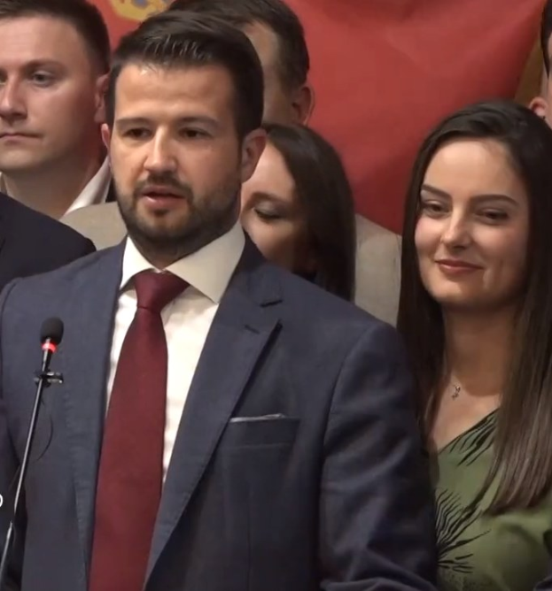 Ein Mann im Anzug spricht in ein Mikrofon an einem Rednerpult, mit einem Banner hinter ihm, zu einer Menge.