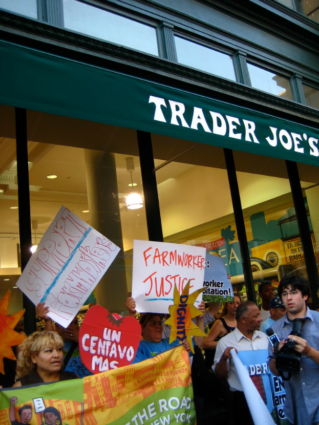 Eine Gruppe von Menschen steht vor einem Geschäft und hält Schilder und Plakate in den Händen, mit einer Säule und Lichtern auf dem Dach im Hintergrund.