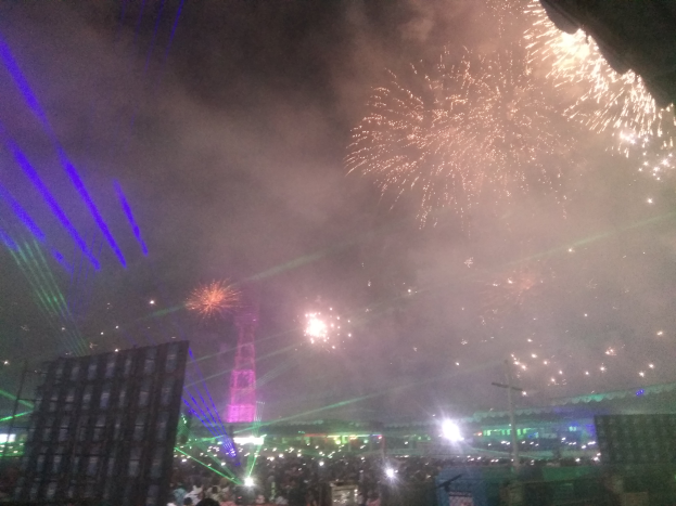 Eine Menschenmenge bei einem Konzert, beleuchtet von Gebäuden und Laternenlichtern, mit farbenfrohen Feuerwerken am Himmel.