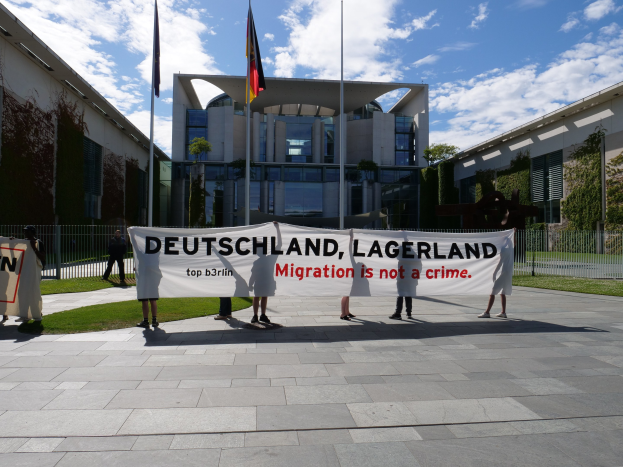 Eine Gruppe von Menschen hält ein Transparent mit der Aufschrift "Deutschland, Lagerland Migration ist kein Verbrechen" vor einem Gebäude mit Fenstern, umgeben von Gras, einem Metallzaun, Pflanzen, Bäumen und Fahnenmasten vor einem bewölkten Himmel.