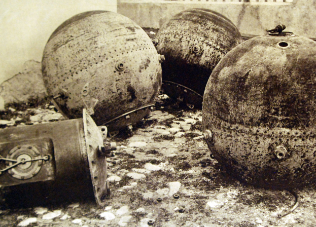 Drei große Metallbomben stehen vor einer Wand mit einem Fenster im Hintergrund, dargestellt in Schwarz-Weiß.