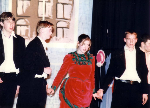 Eine Frau in einem roten Kleid steht in der Mitte, flankiert von Männern in schwarzen Mänteln auf beiden Seiten.