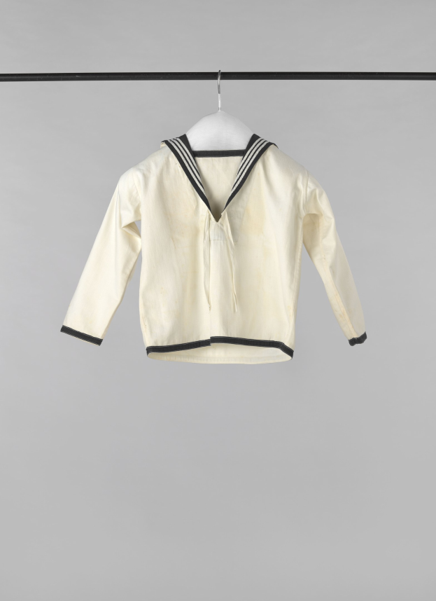 Eine weiße Matrosenjacke mit Kragen, langen Ärmeln und Knopfleiste, die an einer Wäscheleine gegen einen weißen Hintergrund hängt, aus leichtem Rippenstoff hergestellt.