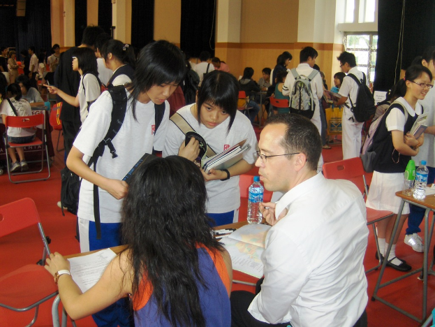 Ein Klassenzimmer mit mehreren Mädchen in weißen T-Shirts, die Bücher in der Hand halten, einem Mann und einer Frau, die im Vordergrund sitzen und diskutieren, und einer gelben Wand mit einem Glasfenster im Hintergrund.